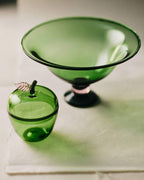 La Pomme, Green decorative glass pieces including a bowl and a small apple jar on a soft background.