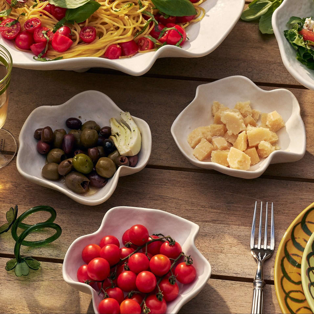 A styled outdoor dining table featuring a large white scalloped platter of spaghetti with cherry tomatoes and basil, surrounded by smaller white dishes of olives, garlic, and cherry tomatoes, alongside patterned yellow and green plates, glasses of white wine, and a wicker chair in the background.