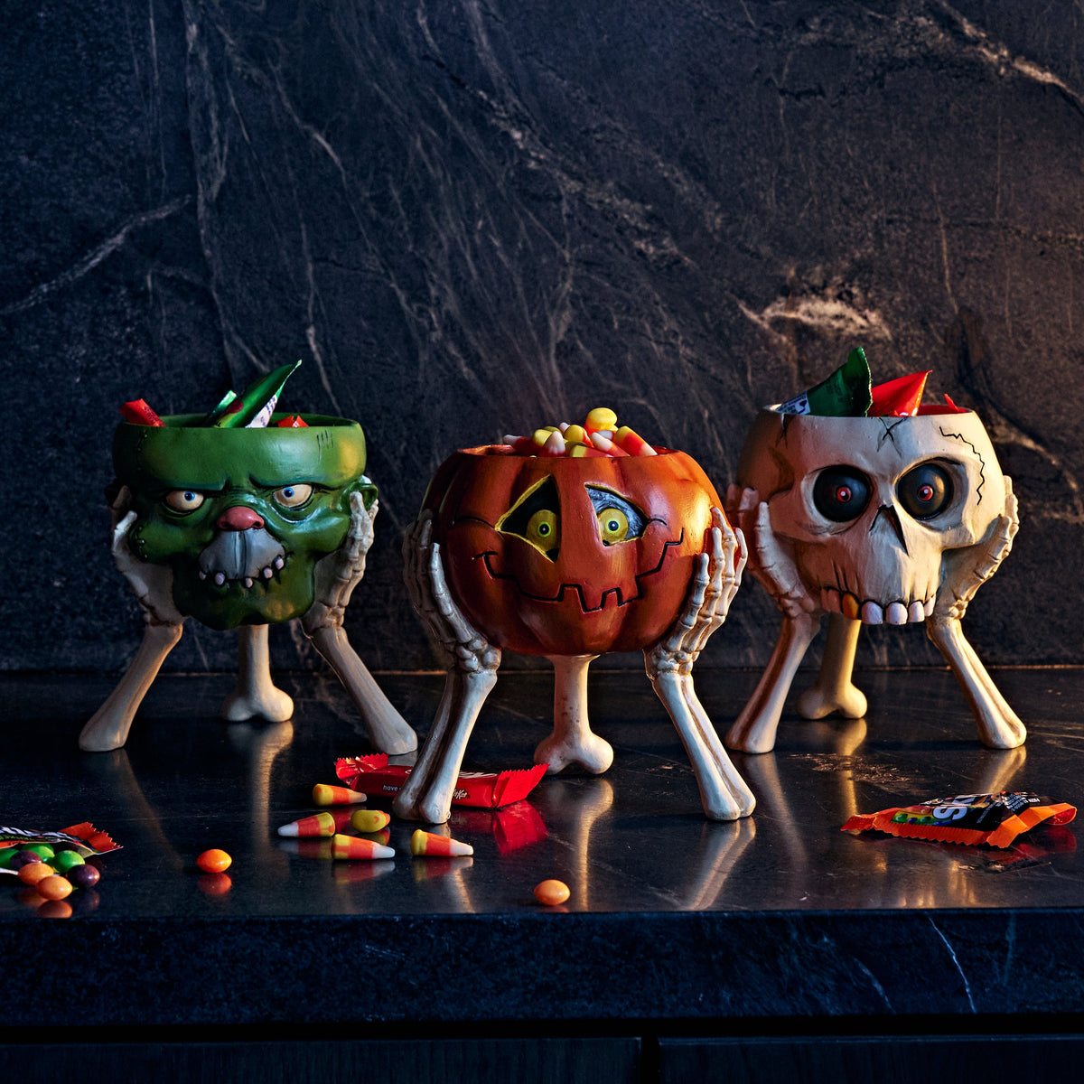 Three Halloween-themed candy bowls shaped like a green monster, a jack-o'-lantern, and a skull, filled with candy and placed on a dark countertop against a dark marble background.