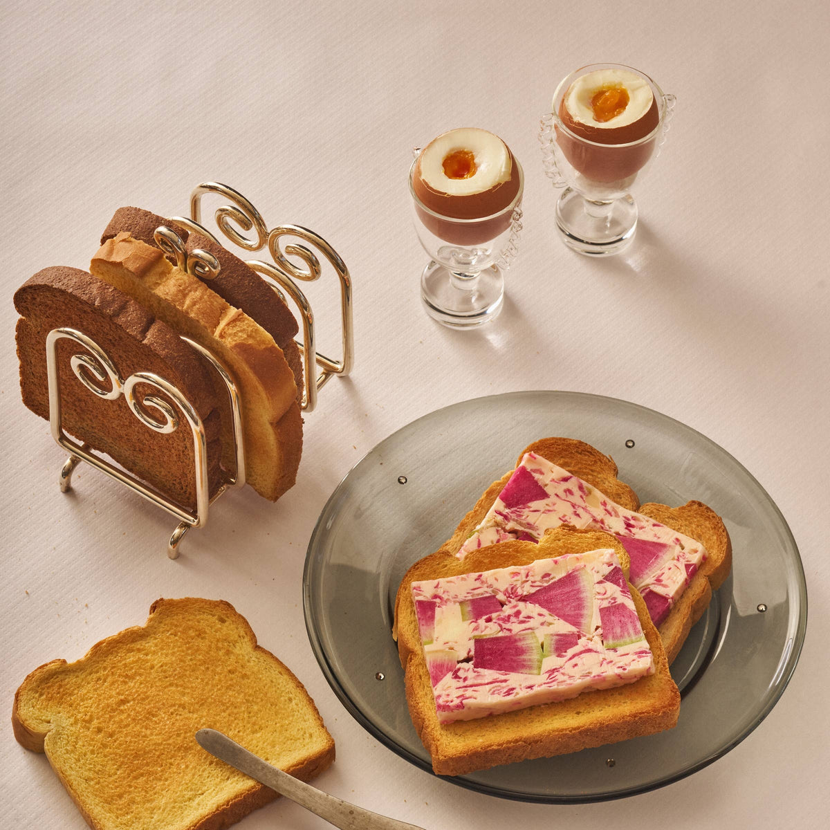 A styled tabletop food scene with several slices of toasted bread, two small glass cups containing browned egg halves, a metal bread rack, and a knife on a pale pink surface.