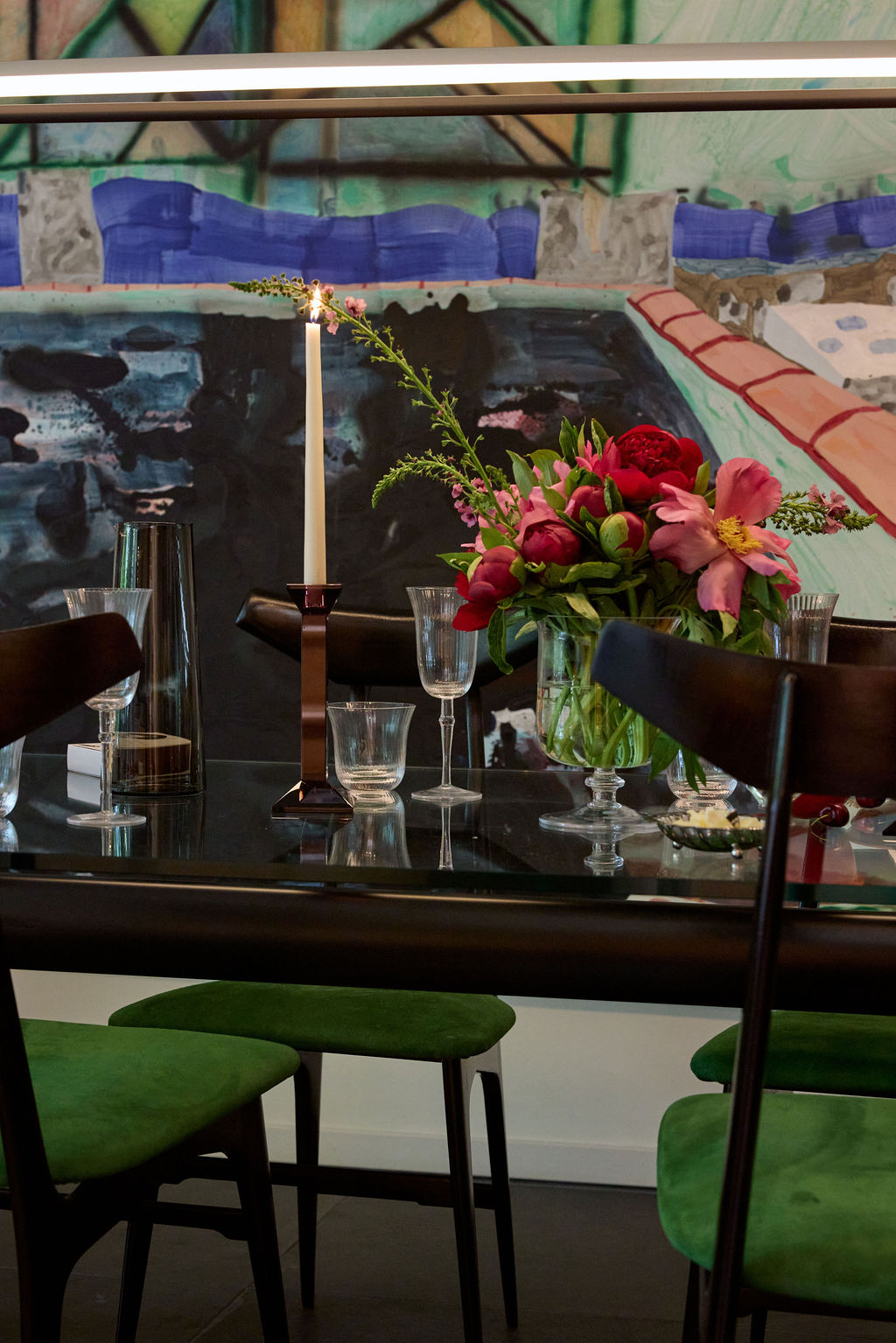 Dining table with a candle, flowers, and green stools in front of a colorful abstract painting.