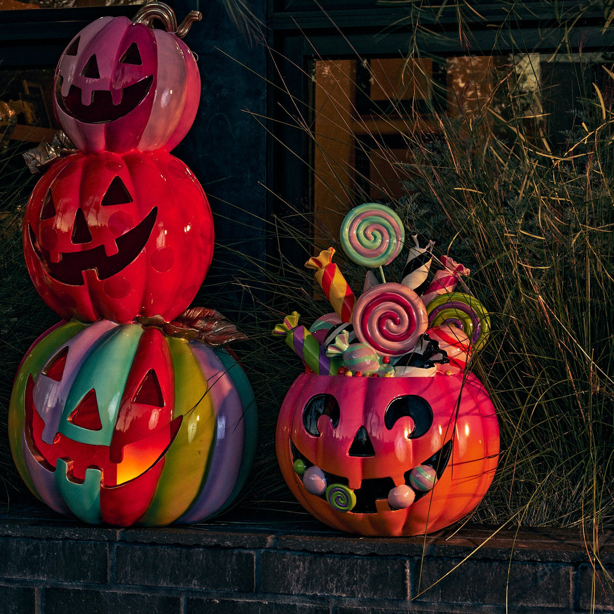 Three decorative jack-o'-lantern pumpkins with candy-filled tops are arranged on a dark brick ledge outdoors at dusk, with tall grasses and a building window in the background.