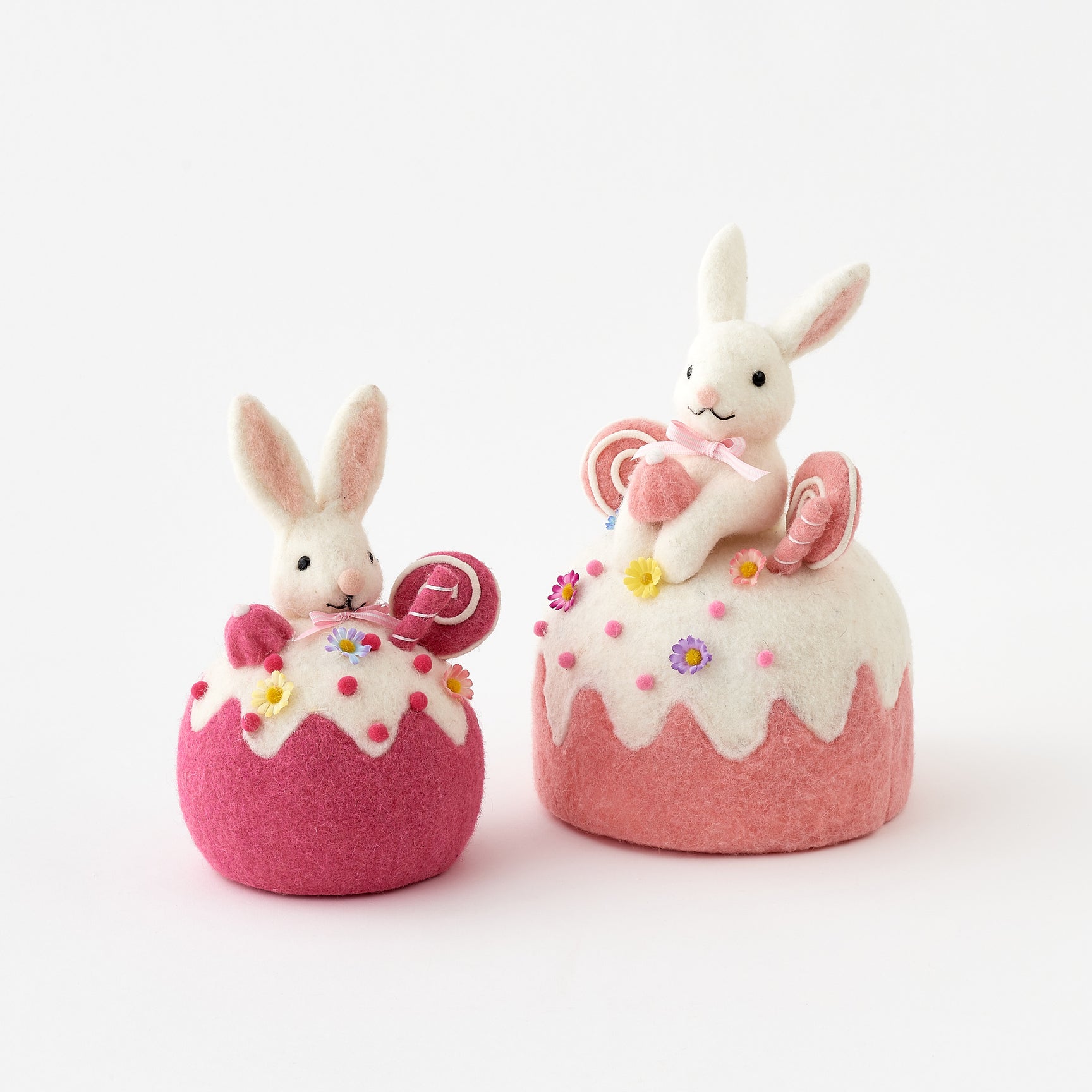 Two felted rabbit toys sitting on pink bases with white frosting and colorful decorations on a white background.