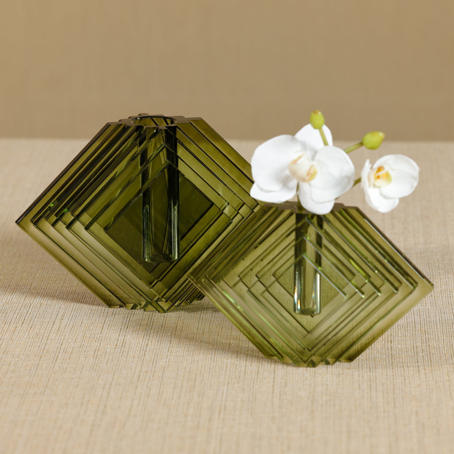 Two green geometric glass vases with a white orchid on a beige surface