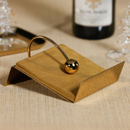 Gold napkin holder on a textured surface with a blurred bottle and glasses in the background