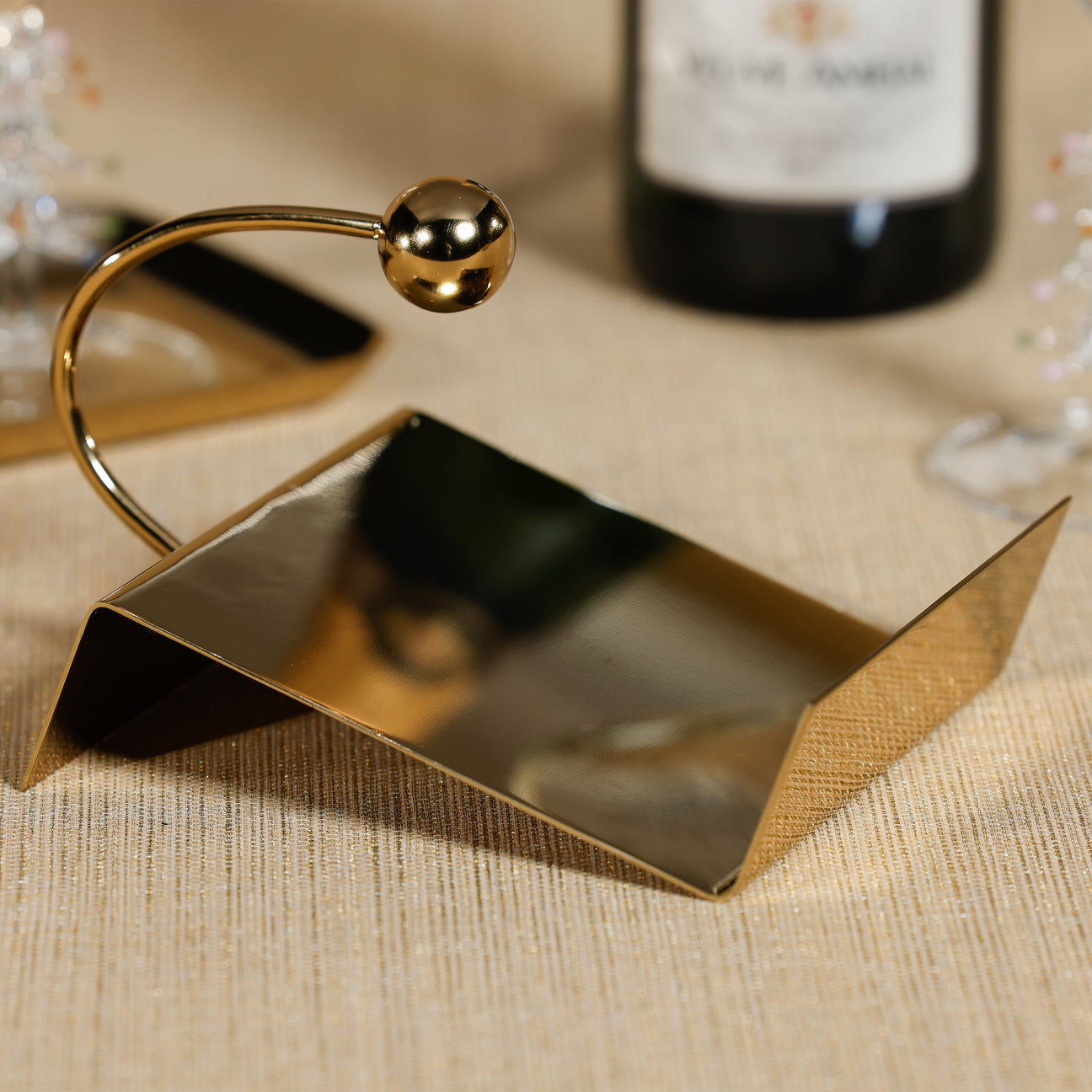 Gold metal napkin holder on a textured surface with a bottle of wine in the background