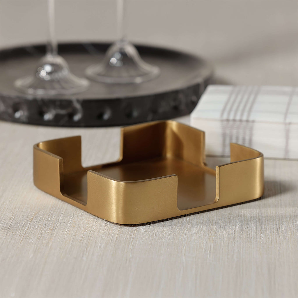 Gold Cocktail Napkin Holder with satin brushed gold finish on a tabletop next to cocktail glasses.