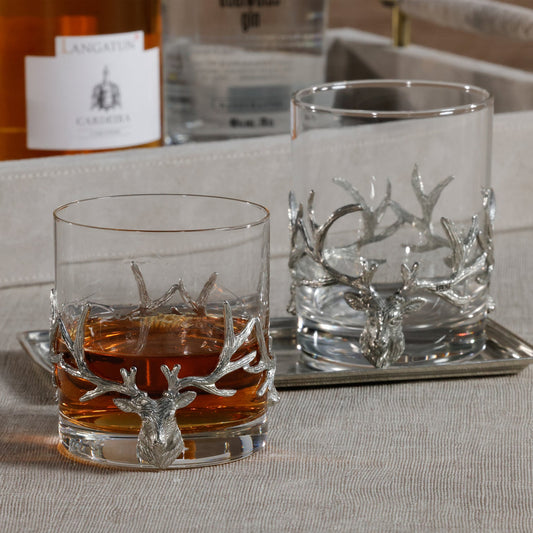 Two whiskey glasses with deer antler designs on a tray, one filled with whiskey.
