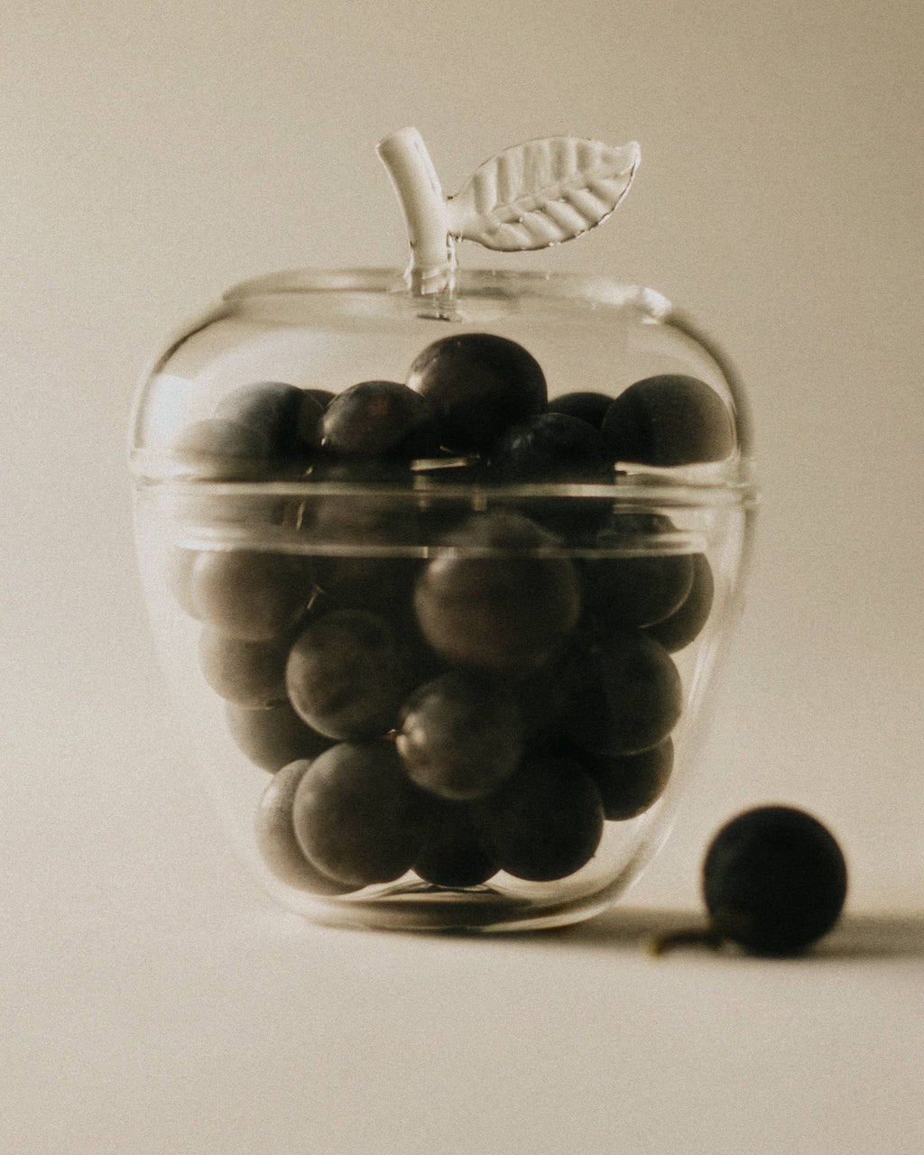 La Pomme, Clear glass apple filled with dark fruit against a soft background, a subtle decor piece.