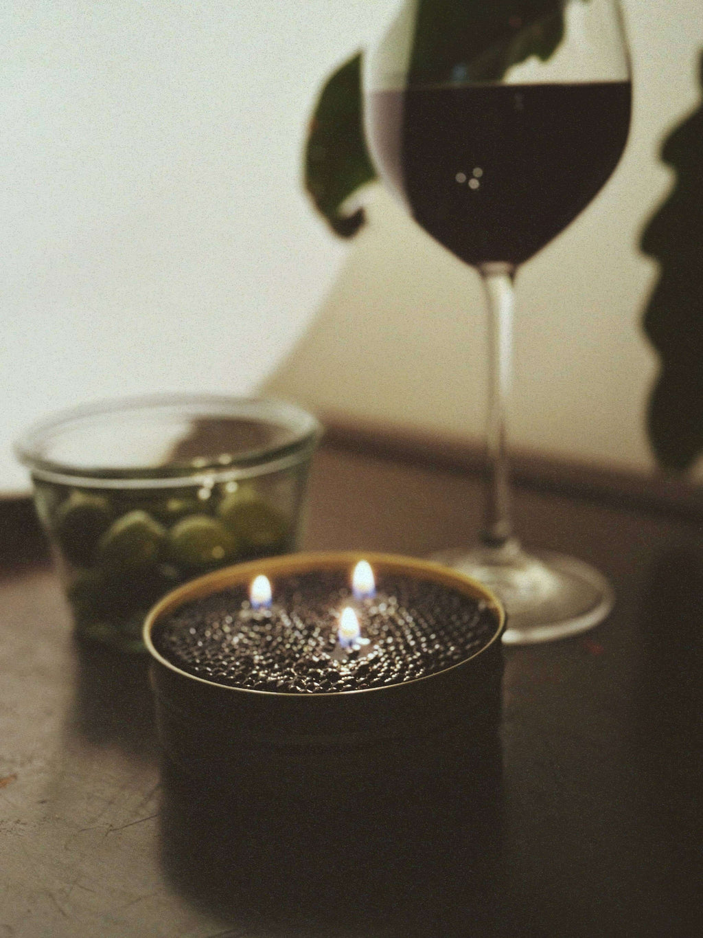 Fancy Caviar Candle in a caviar tin with two flames, alongside a glass of red wine and green olives.
