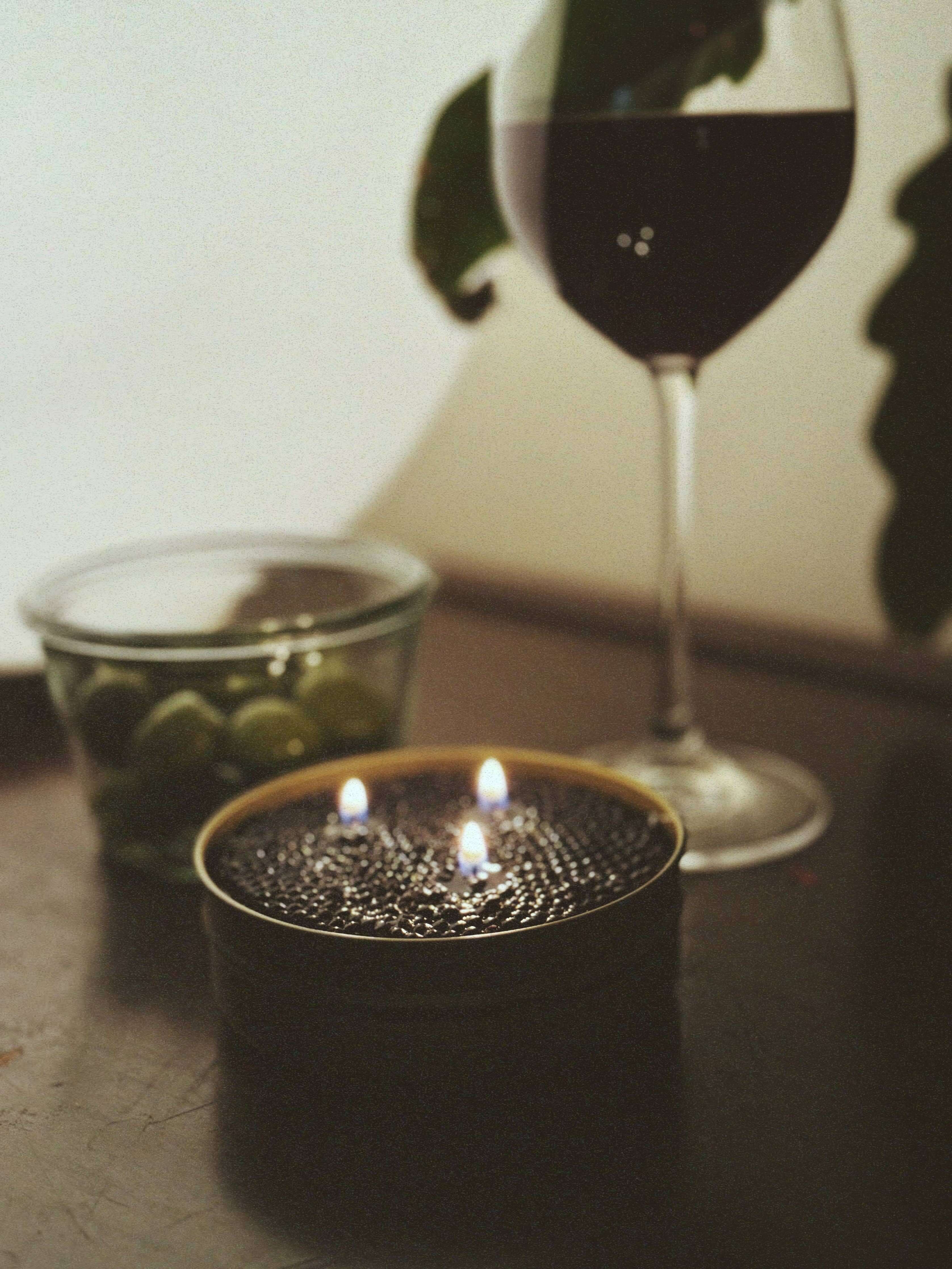 Fancy Caviar Candle in a caviar tin with two flames, alongside a glass of red wine and green olives.